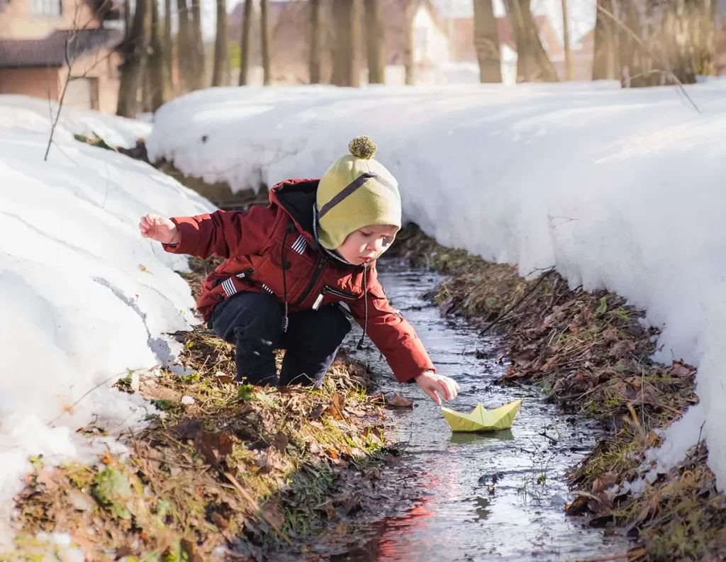 Child in warm clothing floating paper boat on creek in winter snow.