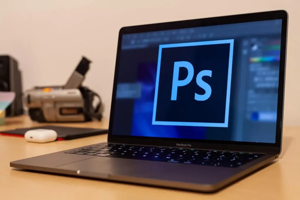 Laptop on desk showing icon of Adobe Photoshop program. A camera is blurry in the background