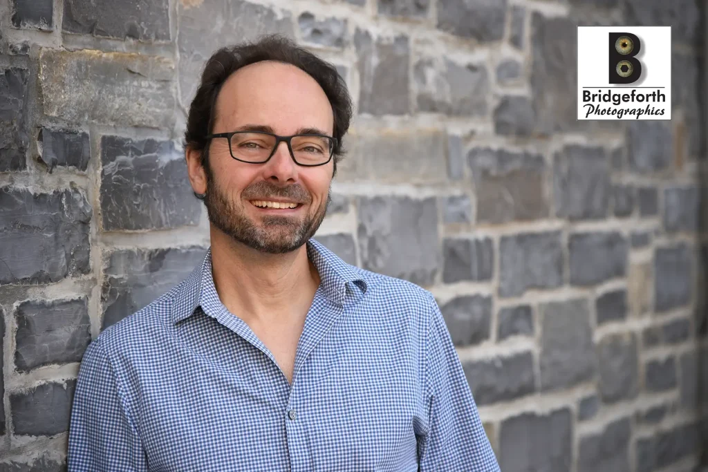 Professional headshot of dark haired man with beard and glasses wearing a blue and white checkered shirt